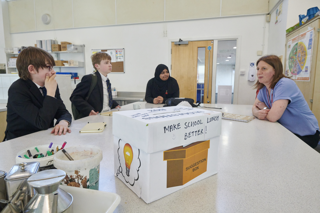 Image shows three secondary school pupils discussing how to make school lunch better with a female teacher.